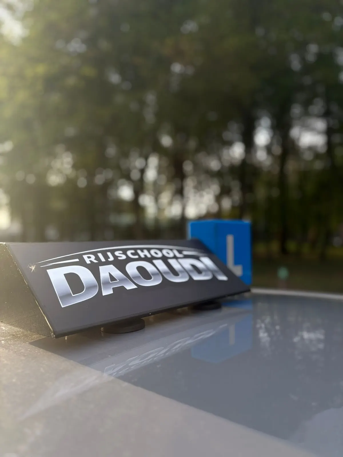 Dakbord met het logo van Rijschool Daoudi en het blauwe L-bord op de lesauto